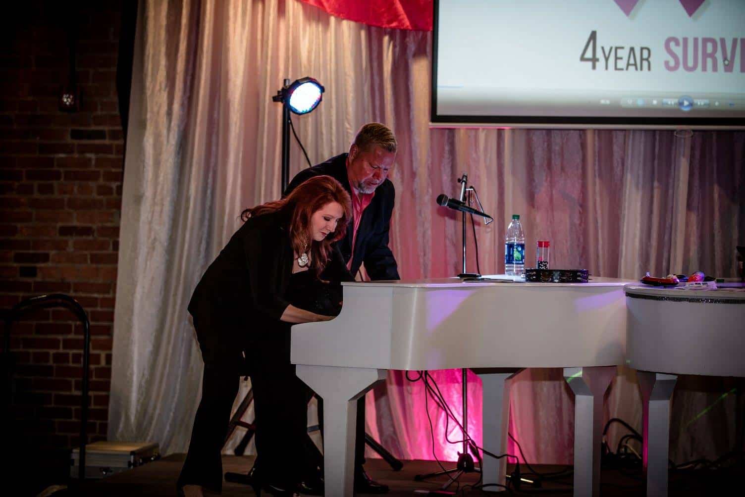 A woman and man playing a white piano on stage at an event.
