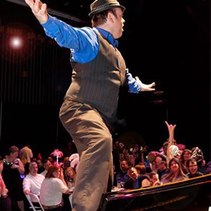 A performer in a hat and vest entertains an audience by standing on a piano during a lively show.