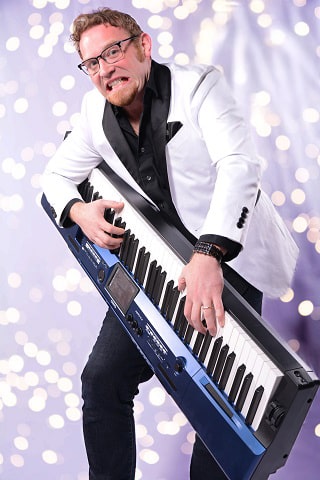 A man in a white jacket enthusiastically plays a keytar against a sparkling background.