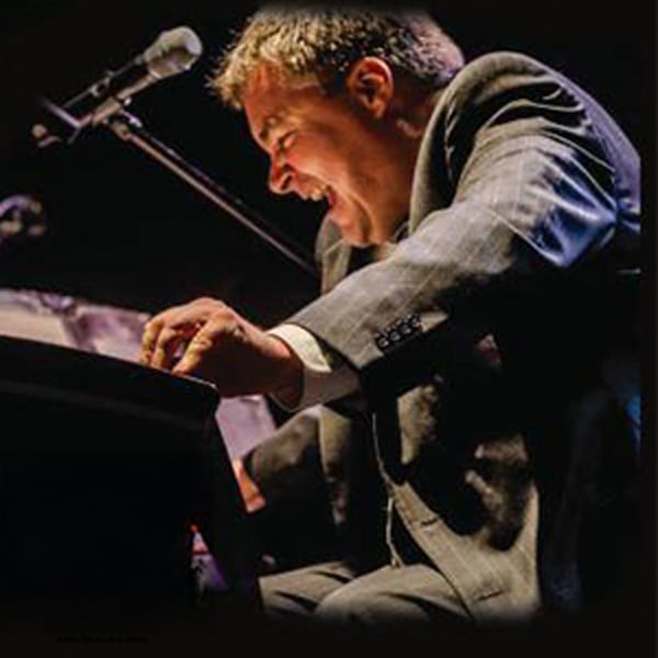 Man in a suit passionately playing piano on stage with a microphone nearby.