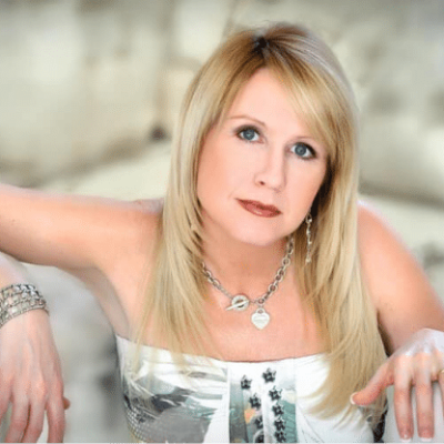 A woman with long blonde hair wearing a necklace and bracelet, sitting indoors.