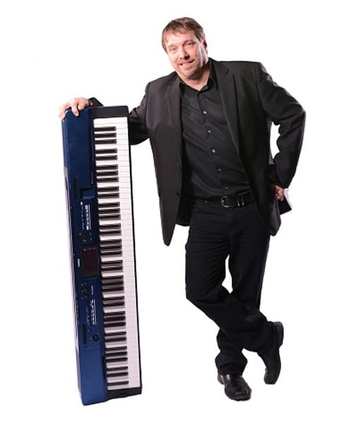 A man in a black suit leans on a blue electronic keyboard against a white background.