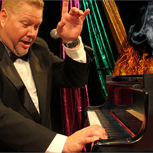 A man in a tuxedo passionately playing a piano with colorful drapes and flames in the background.