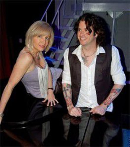 A man and woman smiling and leaning on a piano, with a staircase in the background.