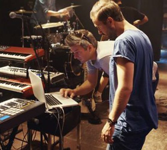 Two men working on a laptop near keyboards and drums on stage.